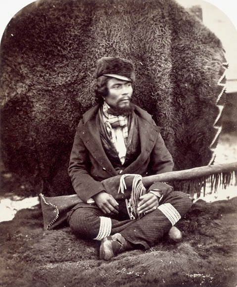 Photographie noir et blanc d'un homme assis sur le sol, jambes croisées. Il tient sur ses genoux un fusil dans son étui.