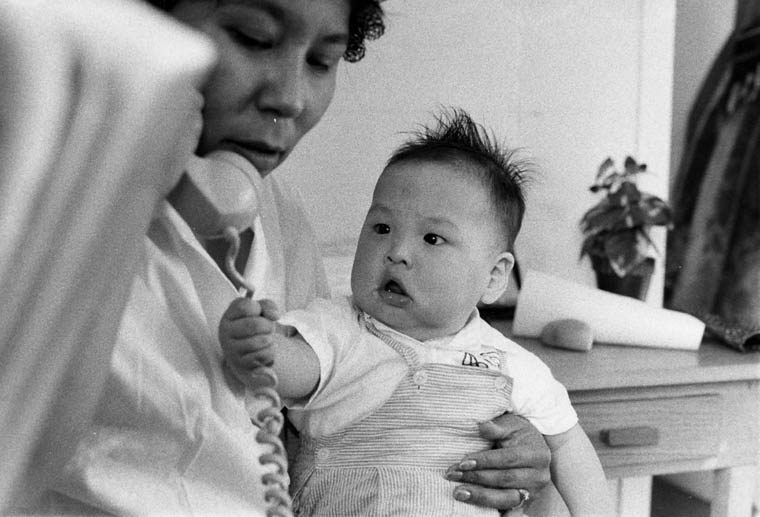 Black-and-white photograph of an Inuit woman talking on the telephone and holding her infant son on her lap, Iqaluit, Nunavut, April 1964