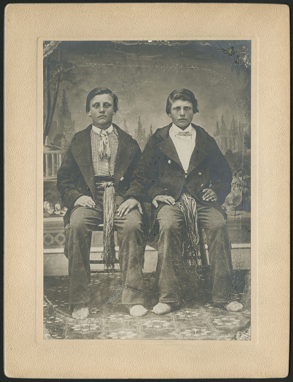 Portrait noir et blanc pris en studio montrant deux garçons assis côte à côte, portant de larges ceintures sous leurs manteaux foncés.