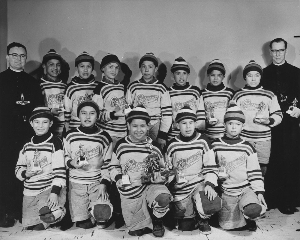 Black and white photo of a boys' hockey team holding trophies. The boys are flanked by two Priests.